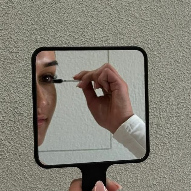 Femme brossant ses cils après un rehaussement, se regardant dans un miroir à main.